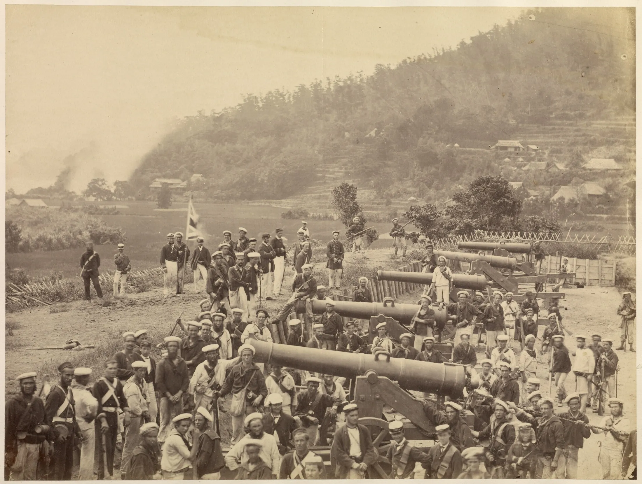 British sailors and marines pose around captured cannon at the Maedamura battery while one man holds a Union Jack above the group.