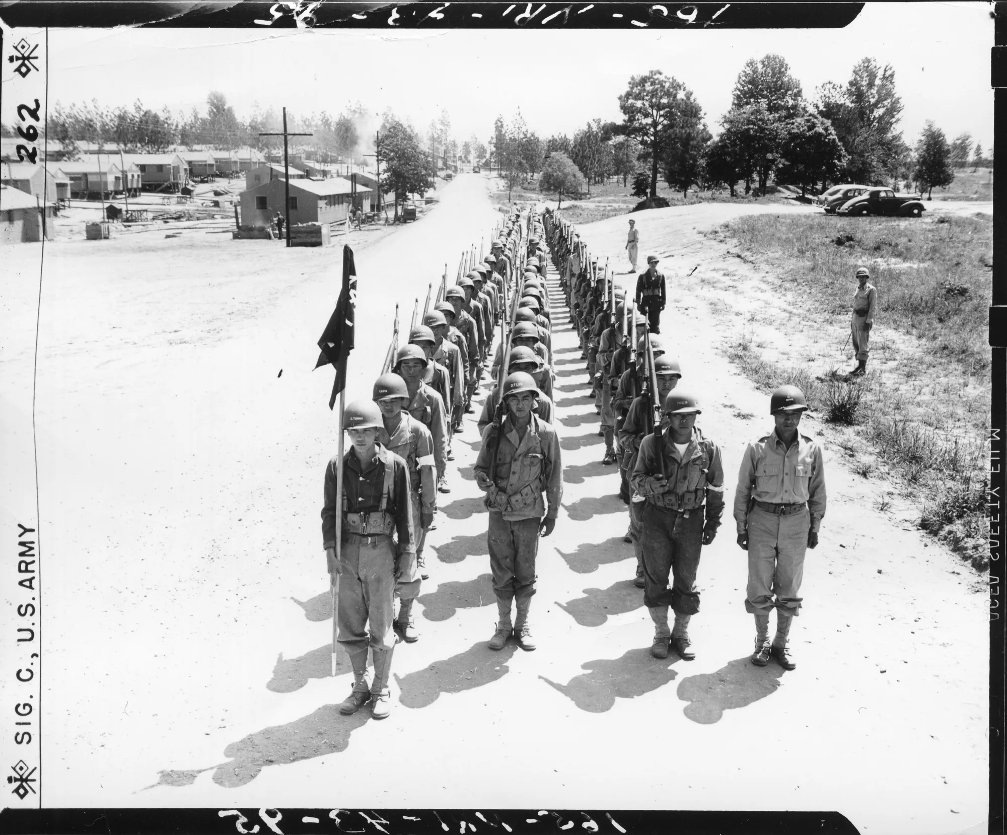 Japanese American soldiers of Company E, 442nd Infantry Regiment stand in long formation at Camp Shelby, Mississippi, with barracks extending behind them.