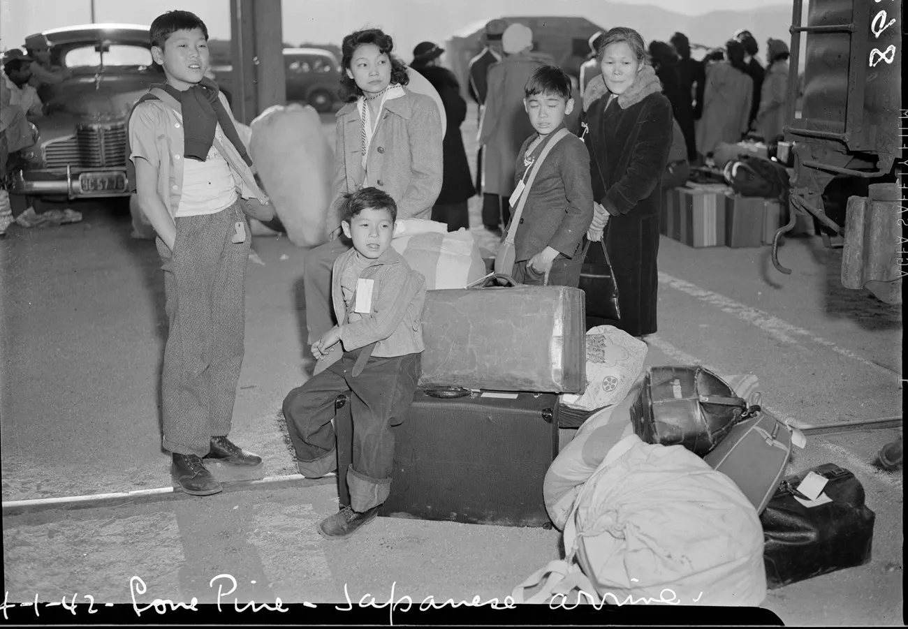 Unidentified Japanese American women and boys wait at Lone Pine with numbered tags, suitcases, bedding, and other people awaiting transport to Manzanar.