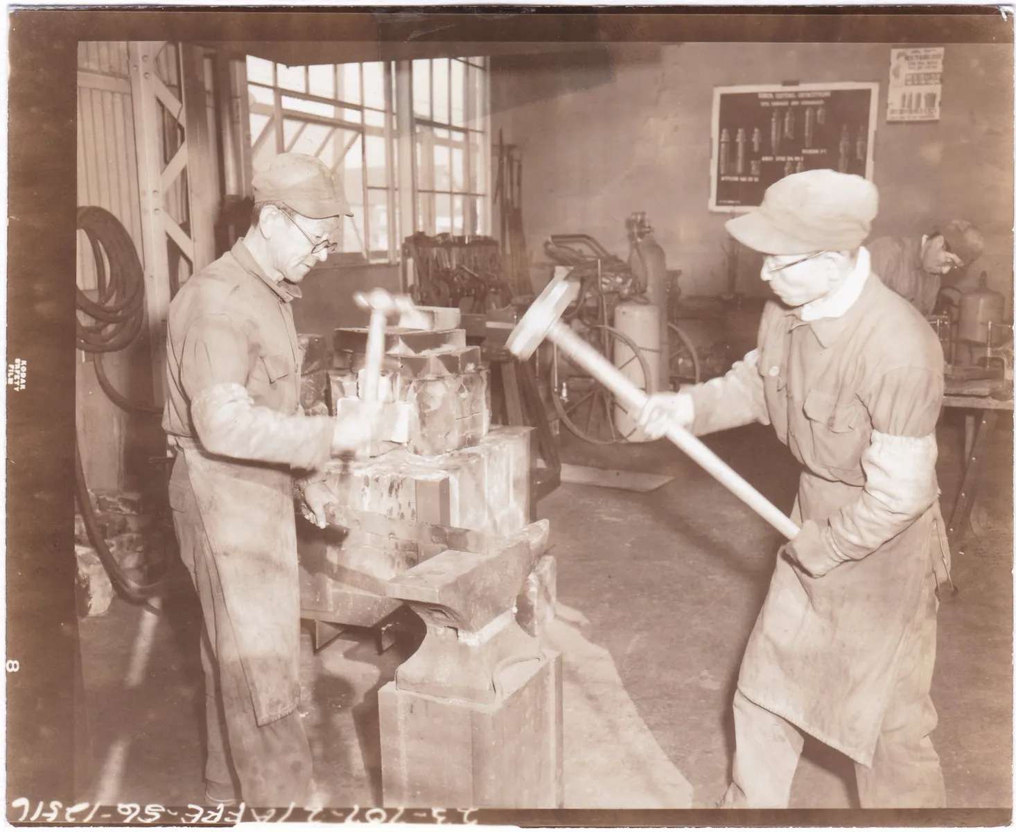 Two Japanese workers in caps and protective aprons swing hammers over an anvil in the blacksmith shop of the U.S. Army ordnance maintenance detachment at Camp Drake, Saitama.