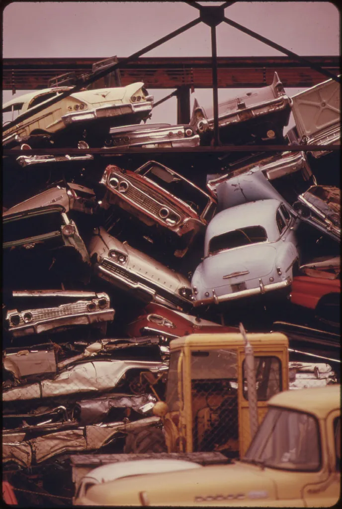 A dense vertical stack of crushed and discarded American automobiles fills a scrap yard, with tail fins, grilles, chrome, and a yellow truck still visible among the wreckage.