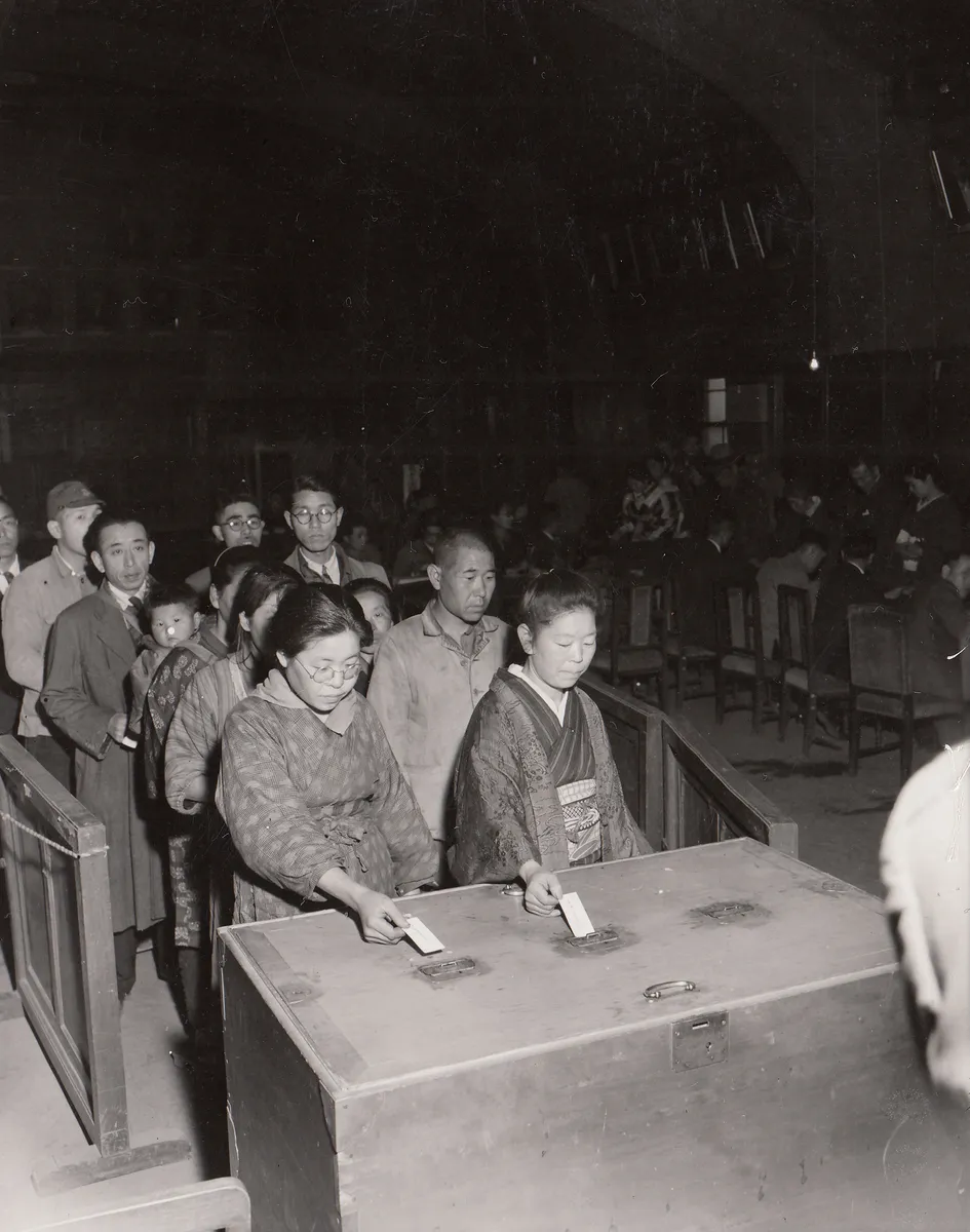 Two Japanese women place paper ballots into a large locked ballot box while men, women, and children wait in a queue inside the Yotsuya Ward office in Tokyo.