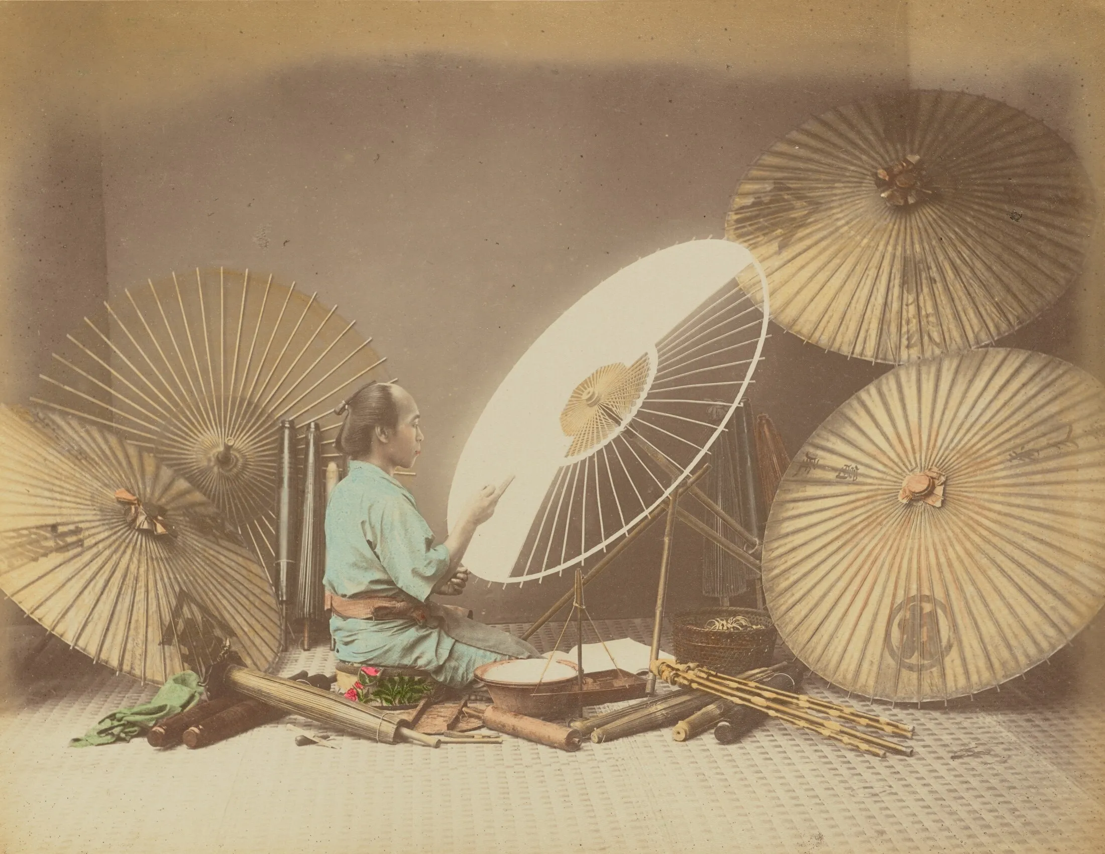 A seated Japanese umbrella maker works on a white paper parasol inside a studio arrangement filled with finished umbrellas, ribs, paper, and tools.