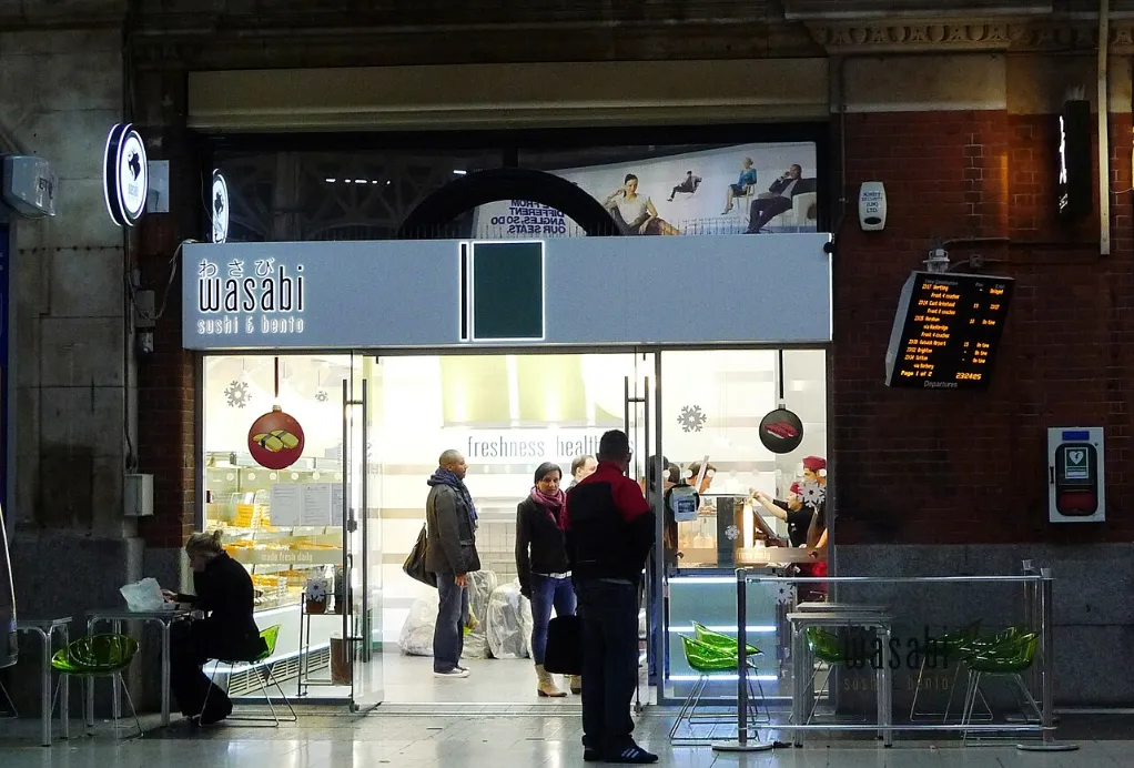 A brightly lit Wasabi sushi and bento takeaway occupies an arched shopfront inside London Victoria station as customers and commuters stand outside.