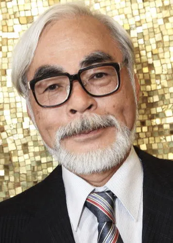 A close portrait of Hayao Miyazaki in black-framed glasses, a dark suit, and a striped tie against a gold mosaic wall at the Venice Film Festival.