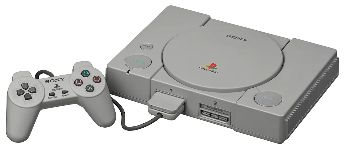 A grey first-model Sony PlayStation sits on a white ground with a wired controller connected, a memory card inserted, and a second memory-card port visible.